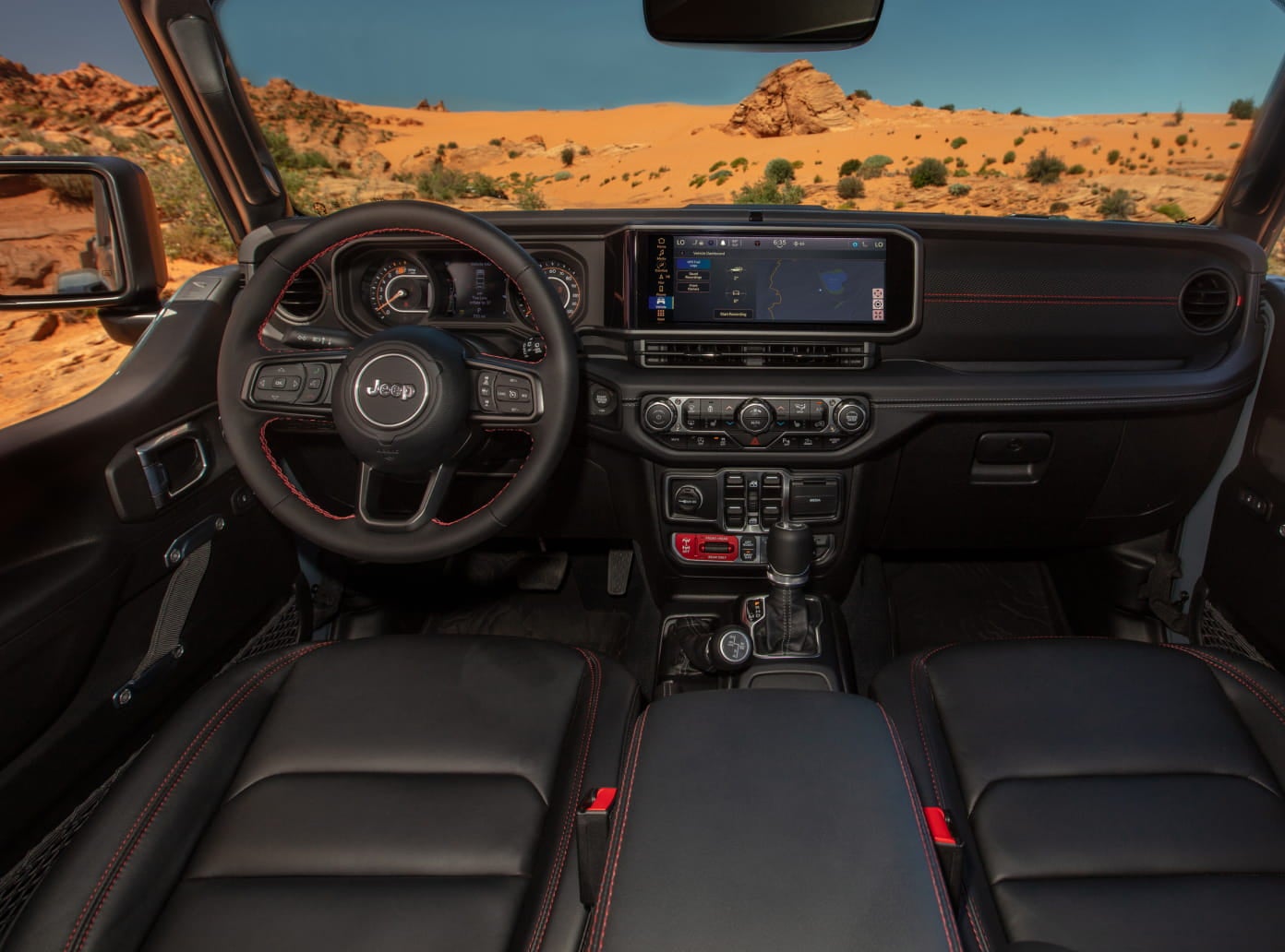 Jeep Wrangler interior with black seats, red stitching, touchscreen display, and a desert landscape visible through the windshield.