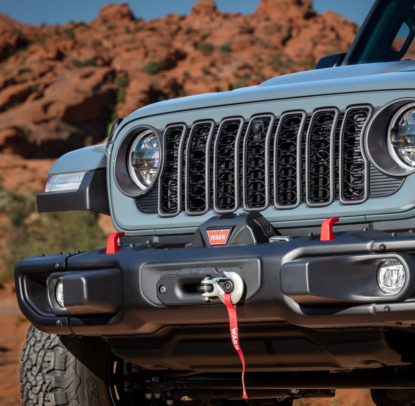 Close-up of a light blue Jeep Wrangler front end with seven-slot grille, round headlights, and integrated winch.