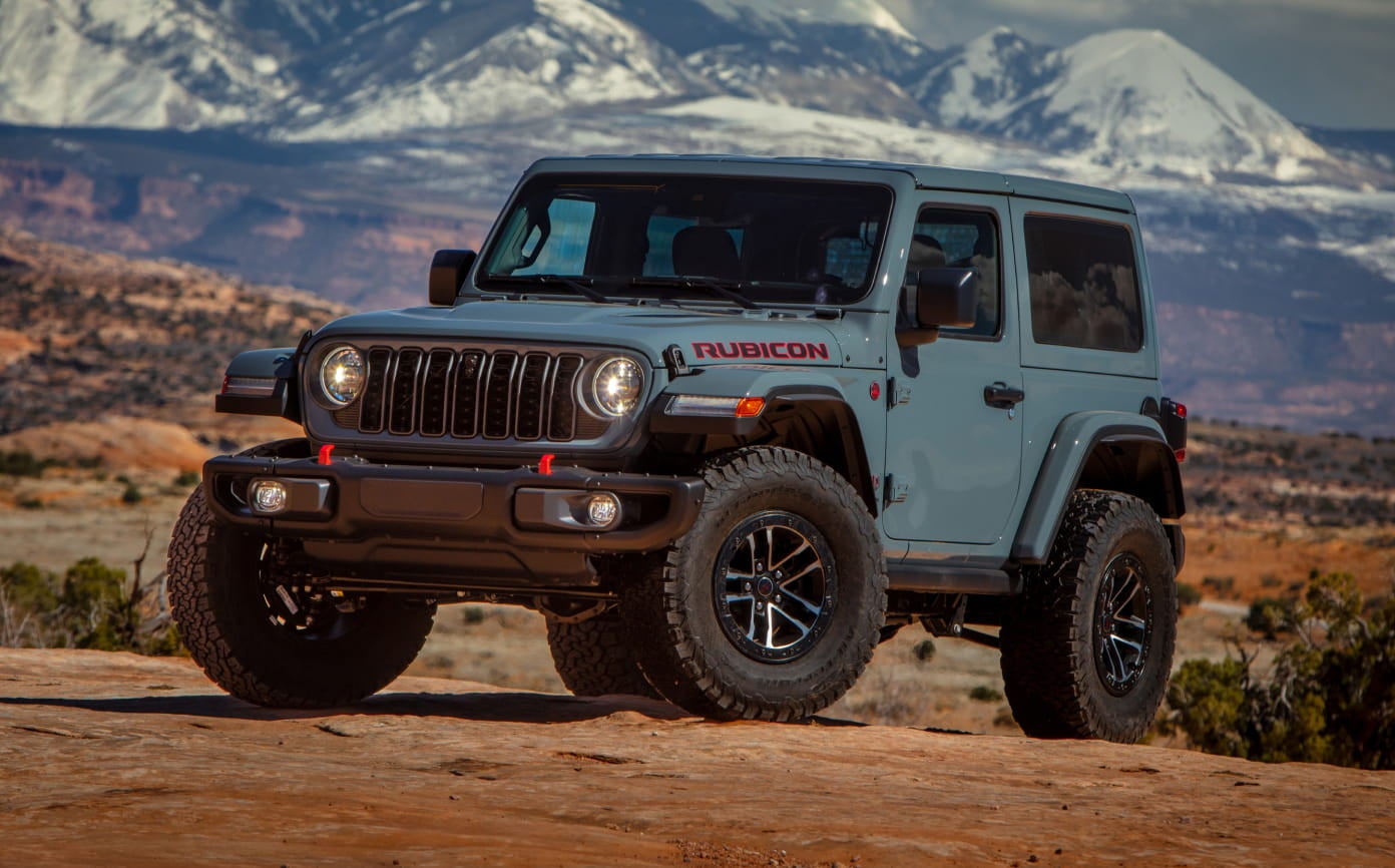 Light blue two-door Jeep Wrangler Rubicon parked on rocky terrain with snow-covered mountains in the background.