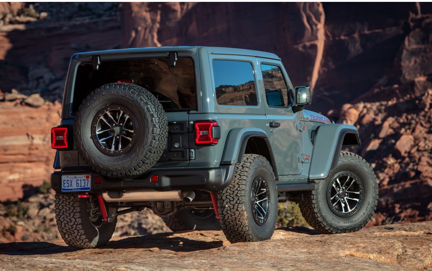 Rear three-quarter view of a light blue two-door Jeep Wrangler Rubicon parked on rocky desert terrain.
